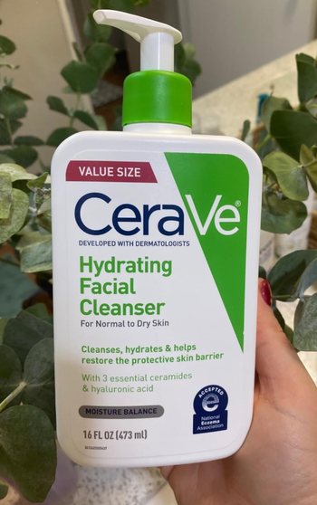 A hand holding a bottle of CeraVe Hydrating Facial Cleanser for normal to dry skin, in a 16 fl oz (473 mL) value size.