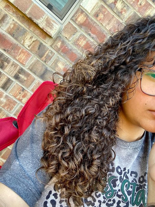 A person with curly hair, wearing glasses and a graphic tee, sitting against a brick wall, partially visible