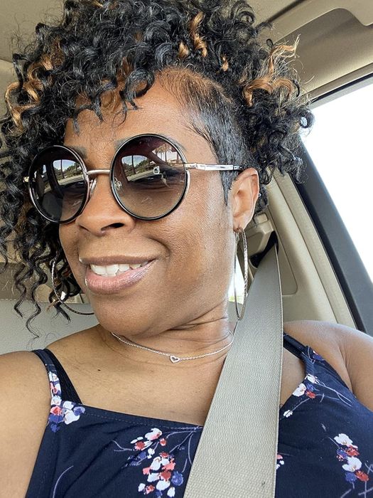 A reviewer with curly hair and oversized round sunglasses taking a selfie in a car, wearing a floral top and seatbelt.