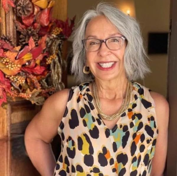 A smiling reviewer with short gray hair and makeup, wearing a sleeveless leopard-print top and a gold necklace, stands in front of a door decorated with a fall-themed wreath.