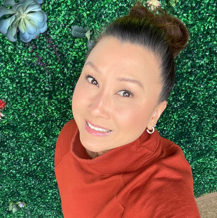 Person smiling, dressed casually in a sweatshirt, surrounded by green foliage.