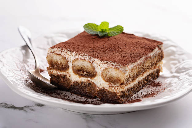 A close-up of a slice of tiramisu on a plate, dusted with cocoa powder and garnished with a mint leaf, accompanied by a spoon.