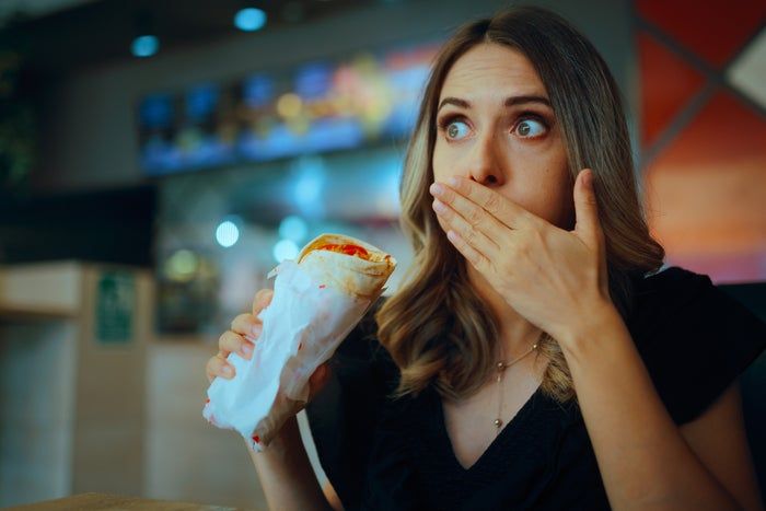 A person in a restaurant holding a wrapped sandwich, covering their mouth with one hand, looking surprised or amused.