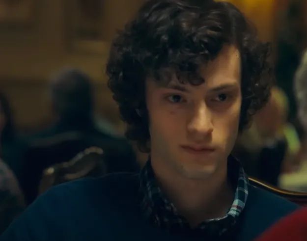 A person with curly hair and wearing a plaid shirt under a sweater, sitting among others, deep in thought