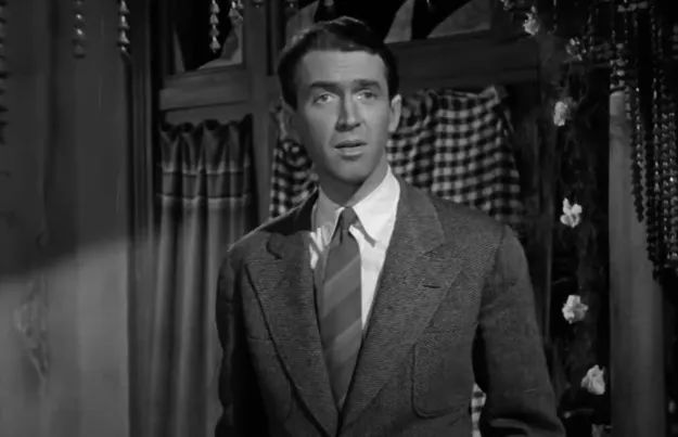A man in a classic vintage suit, standing indoors with a pensive expression, framed by patterned curtains.