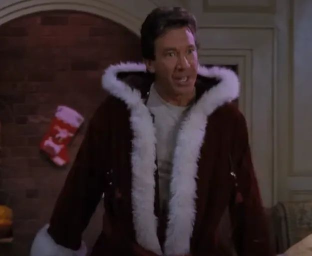 A person dressed in a Santa-like robe with white fur trim, looking either surprised or speaking animatedly. A Christmas stocking hangs on the wall nearby.