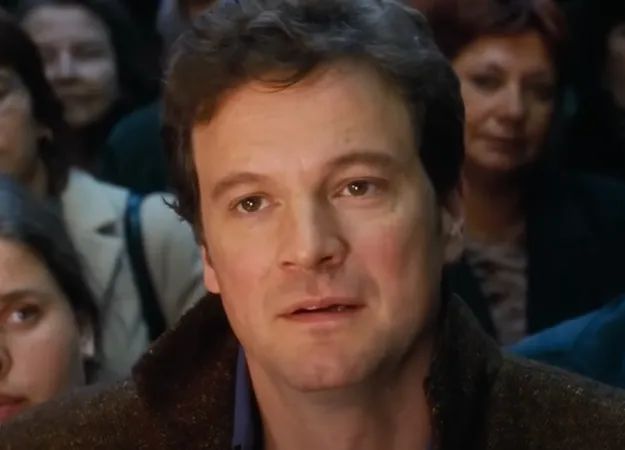 A person stands in a bustling area, staring ahead with focus. He wears a casual jacket and an open-collared shirt, with others blurred in the background