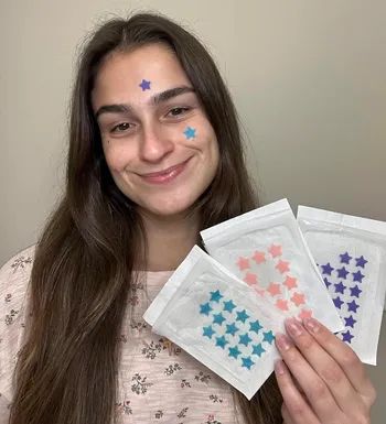A reviewer holding a variety of pimple patches in different colors, with two patches on their face