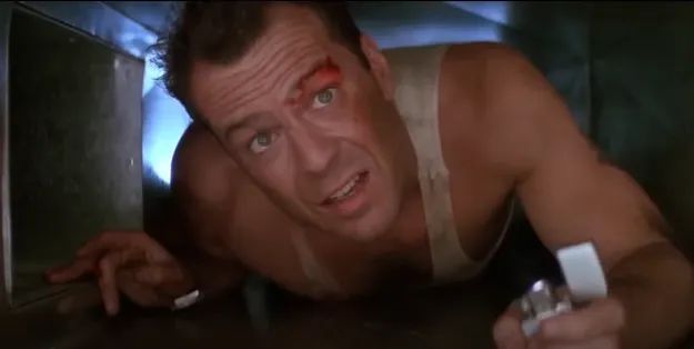 A man in a dusty tank top, crawling through an air duct with a lighter in hand, gazing upwards with a look of determination.
