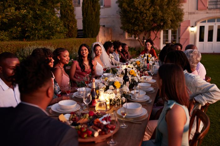 Wedding guests seated at a beautifully arranged outdoor reception table, enjoying a meal and lively conversation. The bride and groom are seen in the center.