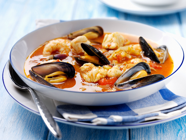 A hearty seafood soup with shrimp, mussels, and fish in a rich broth, served in a white bowl on a table alongside a striped napkin