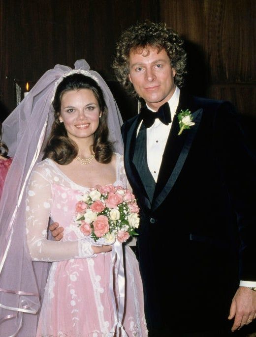 A bride in a long, pink wedding gown with a veil and bouquet, standing beside her groom