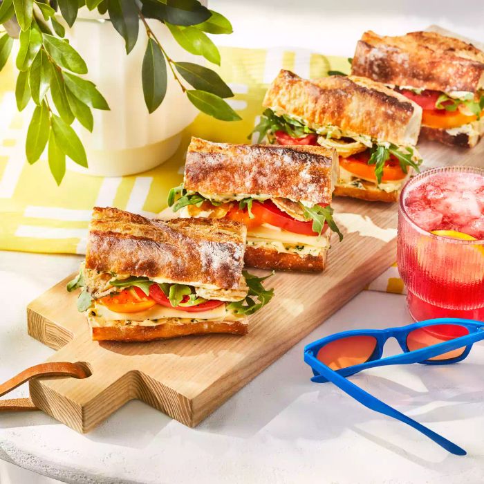 Four tomato sandwiches on a wooden board displayed on a table next to fruit juice, blue sunglasses, and a green plant in a white pot