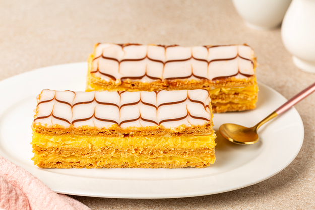 Two layers of mille-feuille pastry with a decorative icing pattern, resting on a plate beside an elegant gold spoon