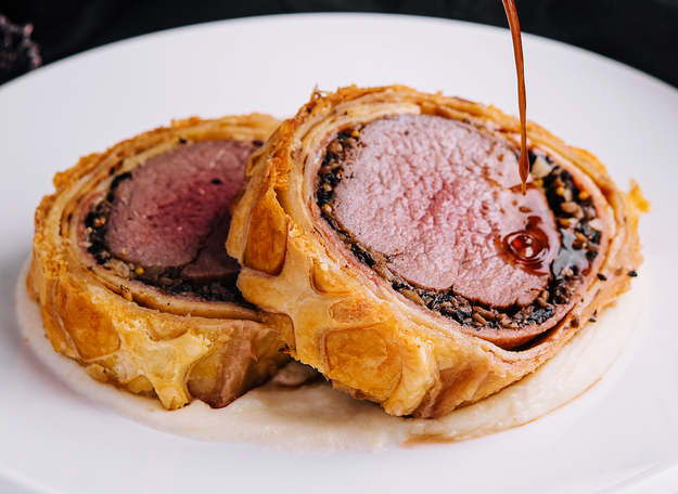 Slices of Beef Wellington on a plate, with sauce drizzled over, highlighting layers of pastry, beef, and duxelles.