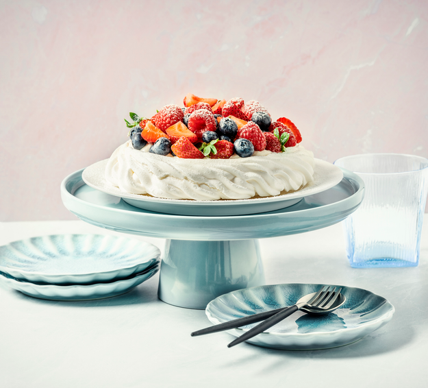 A Pavlova adorned with strawberries, raspberries, and blueberries, placed on a dessert stand and surrounded by plates and cutlery
