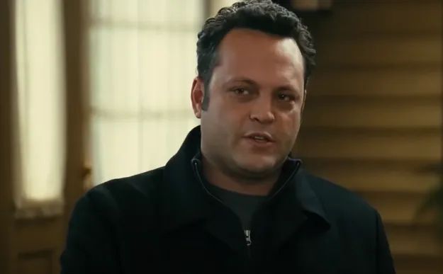 A person wearing a dark jacket, standing indoors, possibly speaking or attentively listening in a laid-back environment.