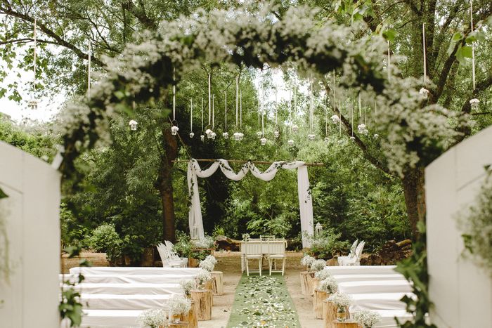 Outdoor wedding venue with floral arch, white seating, and draped ceremony area surrounded by greenery