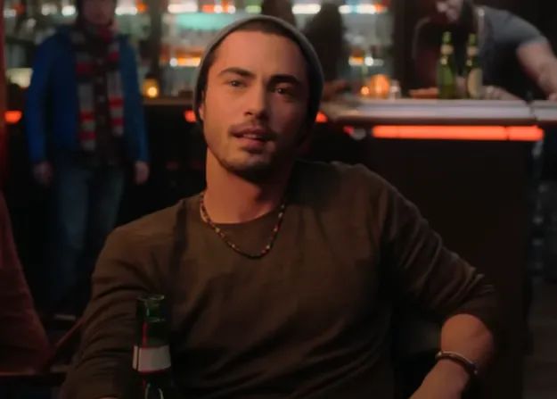 A person in a relaxed atmosphere, wearing a beanie and a necklace, holds a bottle with friends in the background at a bar