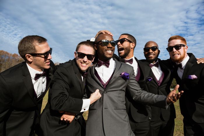 A group of groomsmen in tuxedos and sunglasses happily posing outdoors at a wedding
