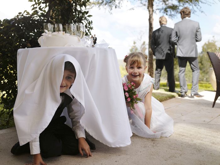 Children are playing hide-and-seek under a wedding table, while two guests in suits stand in the background