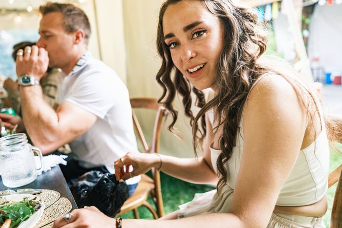 A woman in a casual sleeveless top smiles at the camera while sitting at a dining table with others, reaching to pet a small dog
