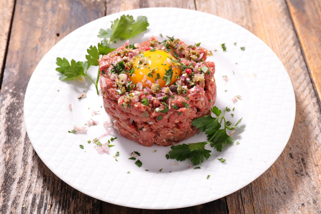 A plate of steak tartare, topped with a raw egg yolk and garnished with fresh herbs and parsley, resting on a wooden surface