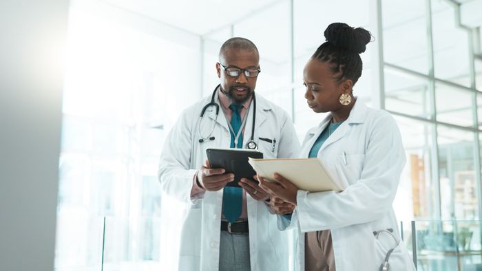 Two doctors, one holding a tablet and the other a folder, discuss in a hospital corridor. They’re both dressed in white lab coats