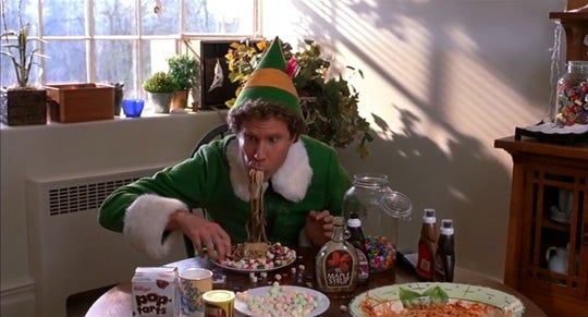 An elf-costumed individual happily enjoying candy-topped spaghetti at a table filled with assorted snacks and syrup