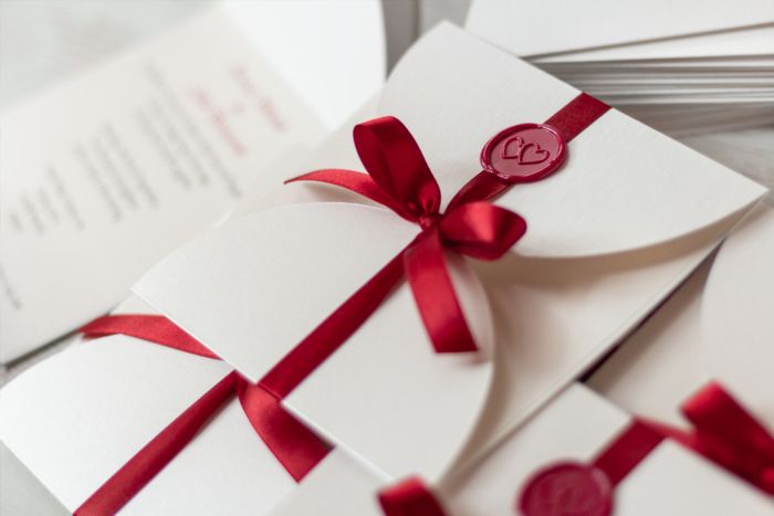 An elegant wedding invitation featuring a red ribbon and heart-shaped seal, displayed alongside other similar invitations