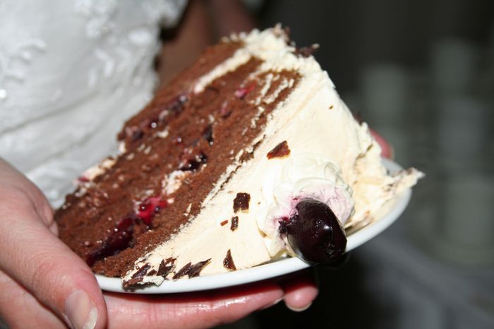 A close-up of a slice of wedding cake on a plate, with chocolate layers, cream frosting, and a cherry on top
