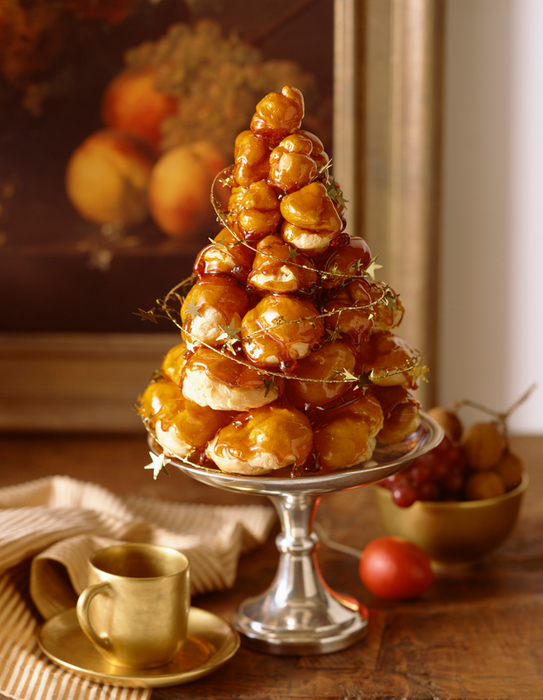 An exquisite croquembouche displayed on a silver stand, drizzled with golden caramel and adorned with decorative sprigs, accompanied by a gold teacup and surrounded by fruit in the backdrop