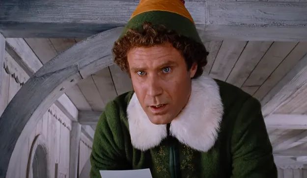 A person in a green outfit with a white fur collar and elf hat, gazing at a piece of paper in astonishment, from a scene in the movie.