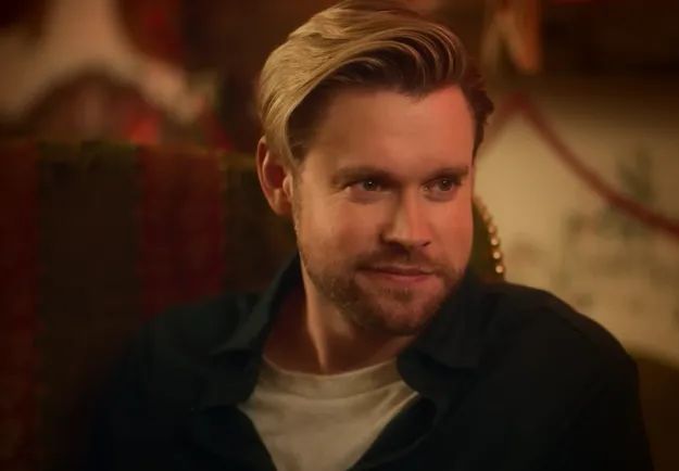 A person with neatly styled hair and a groomed beard, wearing a casual shirt, sitting indoors in a cozy, warm-lit setting.