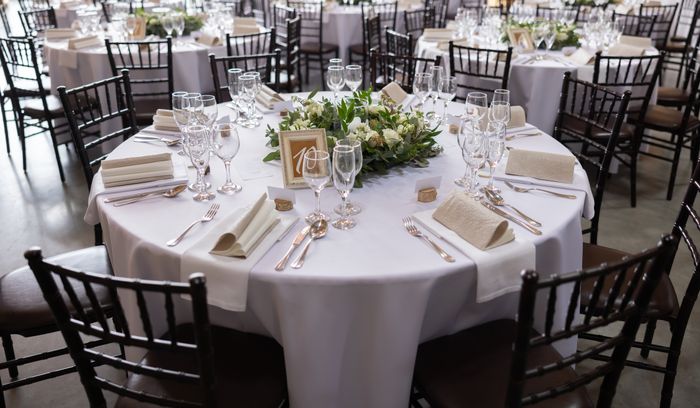An elegant wedding reception table set with white tablecloths, glassware, silverware, and a floral centerpiece. Several tables and chairs surround the setup