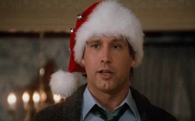 A person in a festive holiday setting, wearing a Santa hat with bells, a suit, and a loosely tied tie