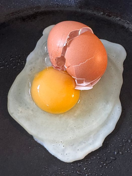 A cracked egg frying in a pan with some of the broken eggshell visible around the yolk.