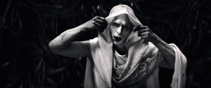 Christian, in full costume, pulls the hood over his head, displaying a dramatic expression and intense makeup in a dark, atmospheric environment.