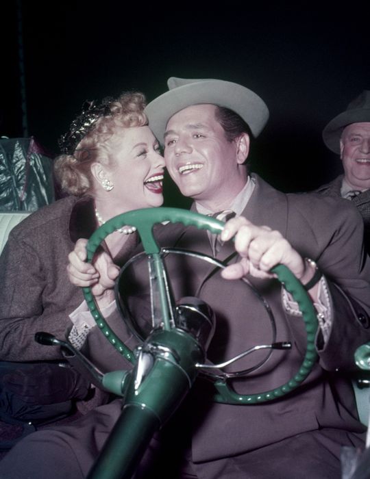 A couple happily poses behind the steering wheel of a vintage car, evoking the charm of classic Hollywood glamour.