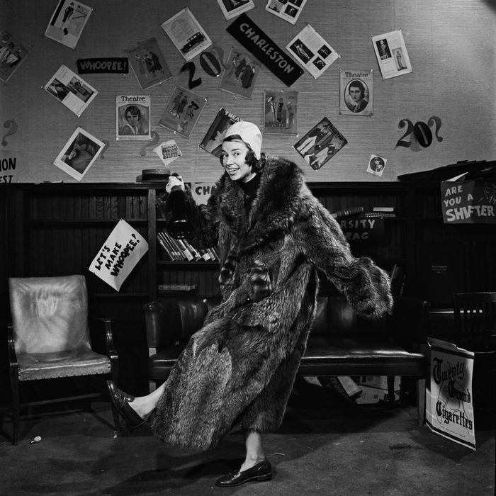 A person in a fur coat strikes a playful pose in a room filled with vintage posters and signs, set in a retro atmosphere with books and a theatrical theme.