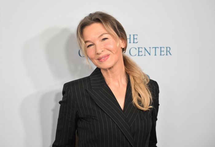 Renée Zellweger smiling at an event, wearing a pinstripe suit and sporting a ponytail hairstyle