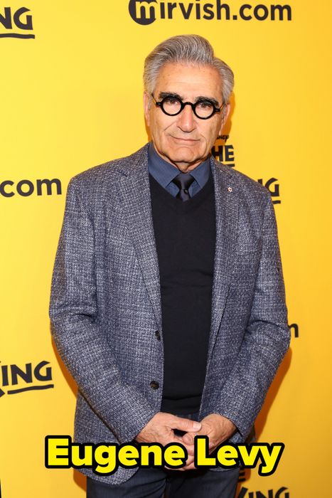 A person in a blue textured blazer and round glasses stands against a yellow backdrop at a celebrity event