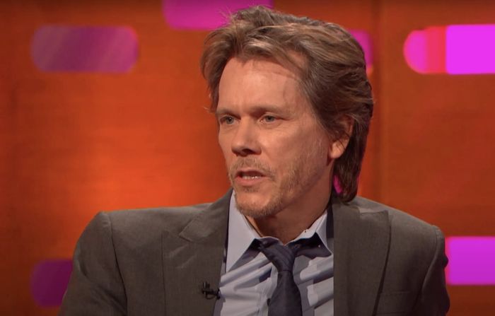 Kevin, dressed in a suit and tie, sits during an interview on a talk show set.