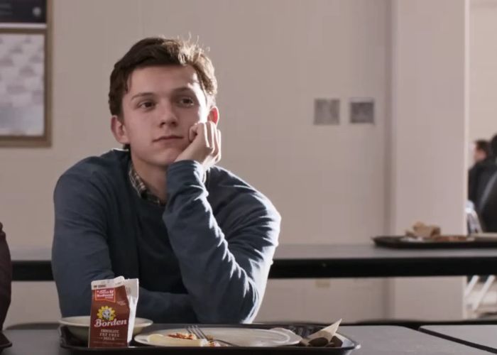 Tom sits in a cafeteria, resting his chin on his hand, looking contemplative. A tray with milk and food is in front of him, dressed in casual attire.