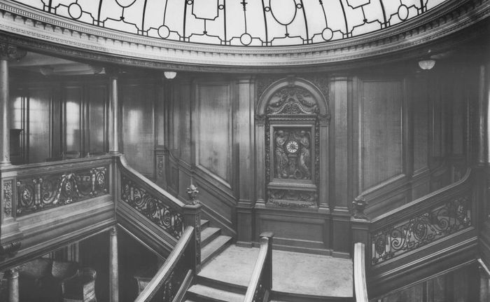 A historic grand staircase with intricate woodwork and ornate railings beneath a decorative glass dome ceiling.