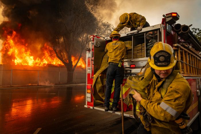 Firefighters in full protective gear urgently work with equipment as a massive blaze rages in the background, with smoke and flames rising high.