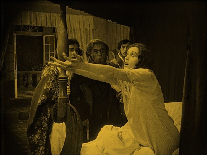 A dramatic moment from a silent film, featuring a woman in distress reaching from a bed, while three people observe from the background