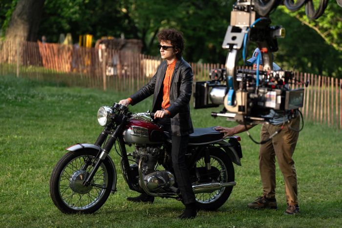 A person wearing sunglasses and a leather jacket sits atop a classic motorcycle during an outdoor film shoot