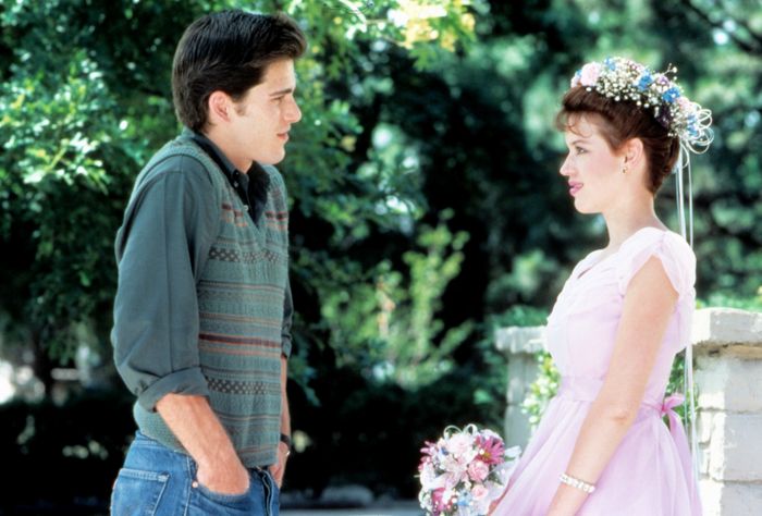 Two people stand outside, facing one another. One is dressed in a sweater over a shirt, while the other wears a floral dress