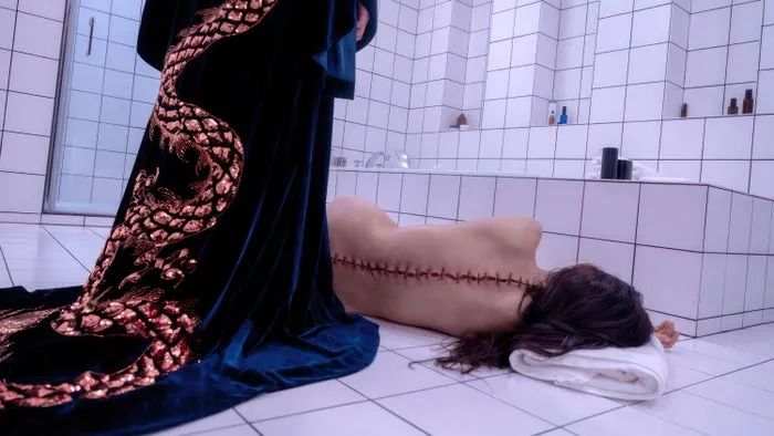A person in an opulent robe with gold detailing stands over another person lying nude on a bathroom floor, their back stitched like a seam.